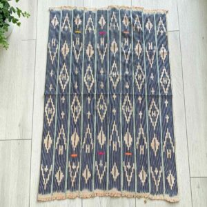 Indigo cotton cloth with columns of pale diamond motifs, laid flat on a timber floor beside a plant