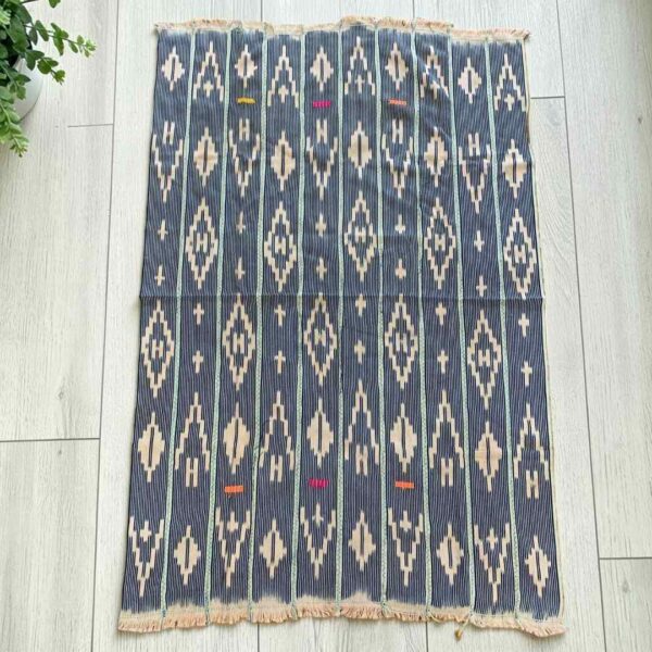 Indigo cotton cloth with columns of pale diamond motifs, laid flat on a timber floor beside a plant