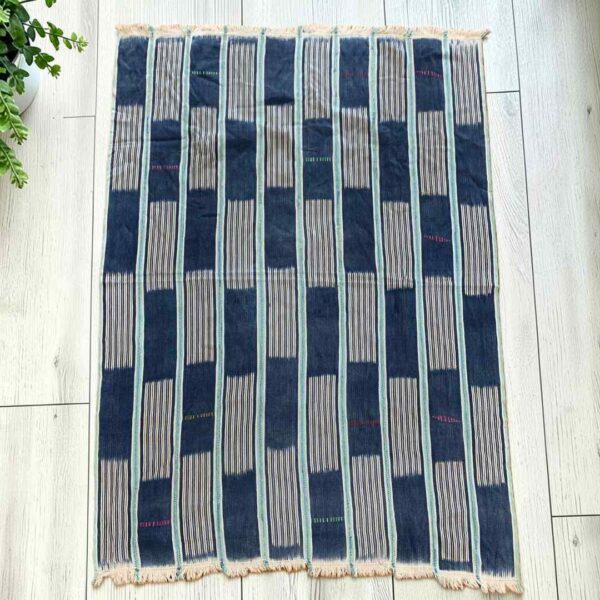 Indigo cotton cloth with solid and pinstriped panels, laid flat on a timber floor beside a plant
