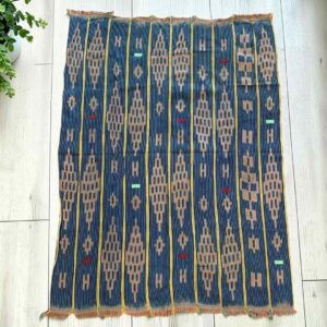 Indigo cotton cloth with chains of warm-toned diamonds, laid flat on a timber floor beside a plant