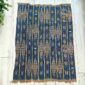 Indigo cotton cloth with chains of warm-toned diamonds, laid flat on a timber floor beside a plant