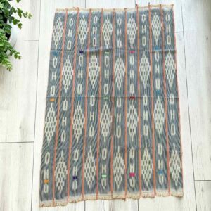 Faded indigo cotton cloth with pale diamond lattice motifs, laid flat on a timber floor beside a plant