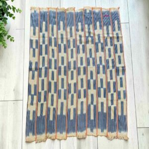 Pale cotton cloth with stepped indigo bars and blocks, laid flat on a timber floor beside a plant