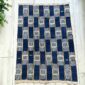 Indigo cotton cloth in a checkerboard of deep and pale blocks, laid flat on a timber floor beside a plant
