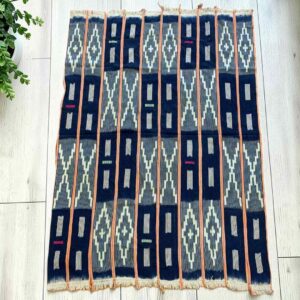 Dark indigo cotton cloth with pale zigzag and window motifs, laid flat on a timber floor beside a plant
