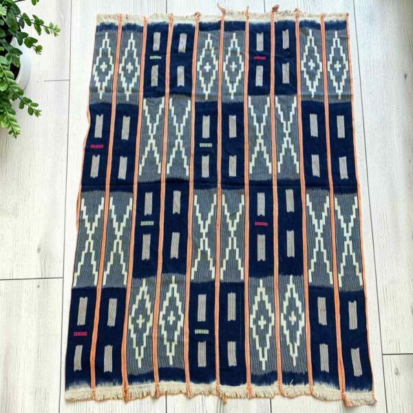 Dark indigo cotton cloth with pale zigzag and window motifs, laid flat on a timber floor beside a plant
