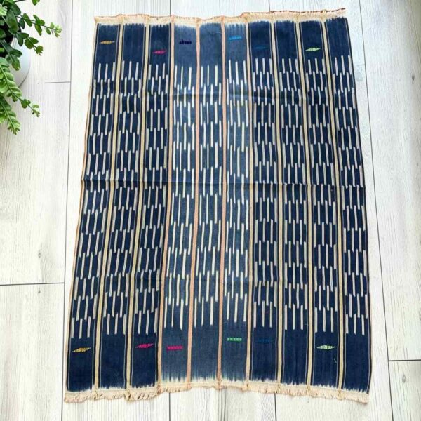 bashiri-indigo-cotton-237-mole-african-textile-handwoven-main Deep blue cotton cloth with columns of pale dashes, laid flat on a timber floor beside a plant