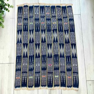 Dark indigo cotton cloth with pale arrow and zigzag motifs, laid flat on a timber floor beside a plant