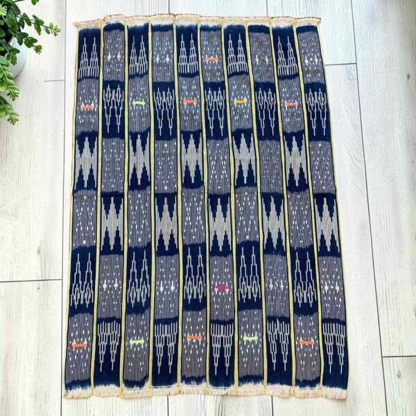 bashiri-indigo-cotton-238-volta-african-cotton-fabric-woven-main Dark indigo cotton cloth with pale arrow and zigzag motifs, laid flat on a timber floor beside a plant