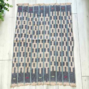 Pale cotton cloth with a stepped pattern of indigo blocks, laid flat on a timber floor beside a plant