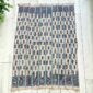 Pale cotton cloth with a stepped pattern of indigo blocks, laid flat on a timber floor beside a plant