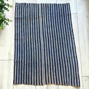 Cotton cloth with fine indigo and pale stripes, laid flat on a timber floor beside a plant