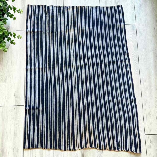 bashiri-indigo-cotton-243-hohoe-handwoven-african-fabric-main Cotton cloth with fine indigo and pale stripes, laid flat on a timber floor beside a plant