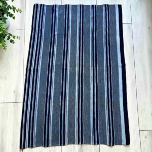 Grey-blue cotton cloth with bands of dark and pale stripes, laid flat on a timber floor beside a plant