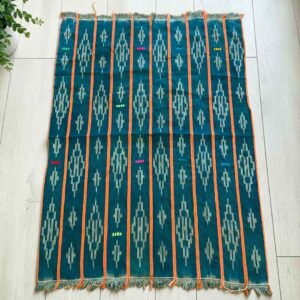 Green-blue cotton cloth with pale diamond motifs, laid flat on a timber floor beside a plant