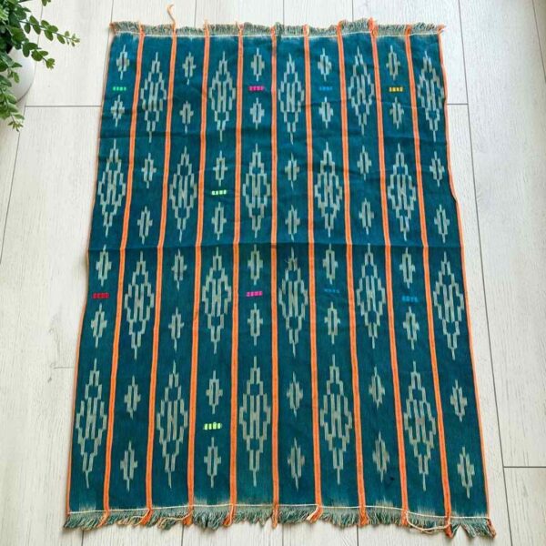 Green-blue cotton cloth with pale diamond motifs, laid flat on a timber floor beside a plant