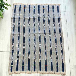 Pale striped cotton cloth with narrow indigo motif columns, laid flat on a timber floor beside a plant