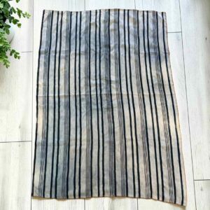 Pale cotton cloth with faded indigo stripes, laid flat on a timber floor beside a plant