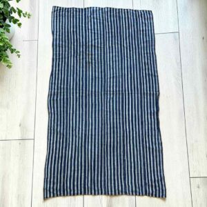 Deep indigo cotton cloth with fine pale pinstripes, laid flat on a timber floor beside a plant