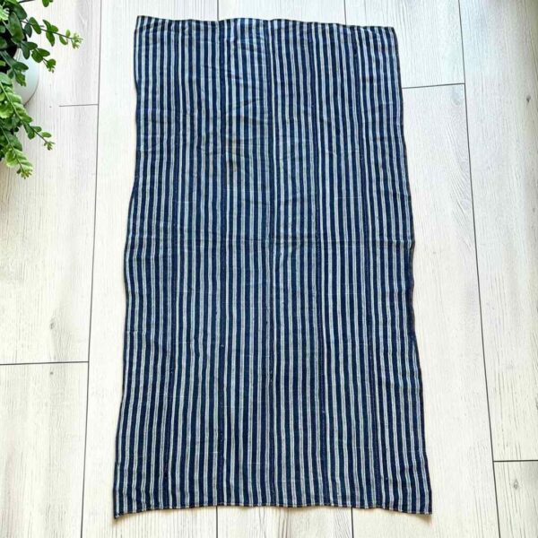 bashiri-indigo-cotton-248-koforidua-african-textile-handwoven-main Deep indigo cotton cloth with fine pale pinstripes, laid flat on a timber floor beside a plant