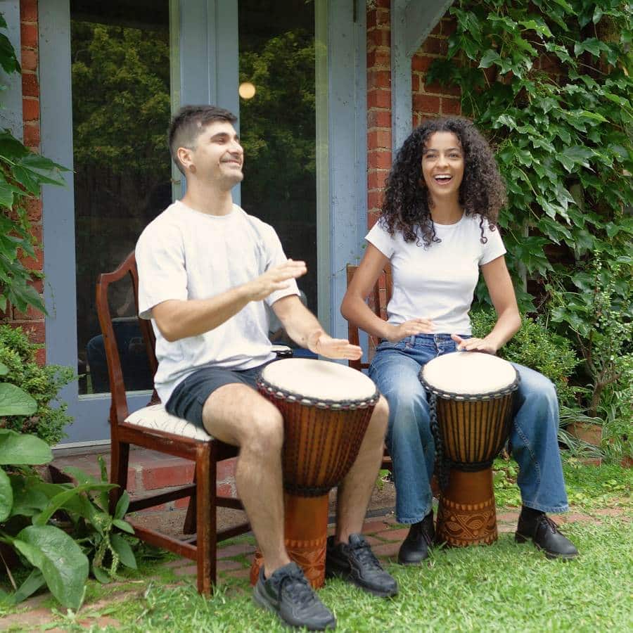 Two people playing Indonesian djembes outdoors, seated in a garden setting, carved mahogany bodies with natural skin heads