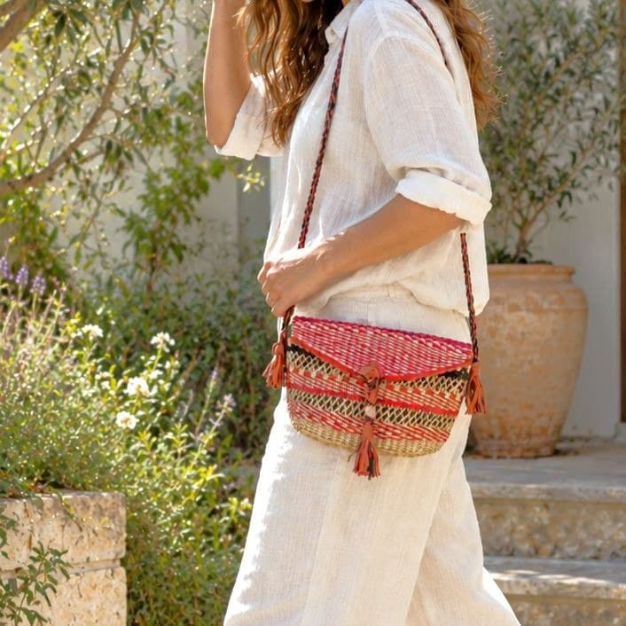 Mini Bolga shoulder bag in red and natural elephant grass with leather tassels and a plaited leather strap, worn at the hip with white linen