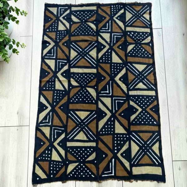 bashiri-mudcloth-medium-161-bustard-african-textile-bogolan-main Medium dark mudcloth with large warm-toned triangles and bow-tie shapes, laid on a timber surface with green leaves