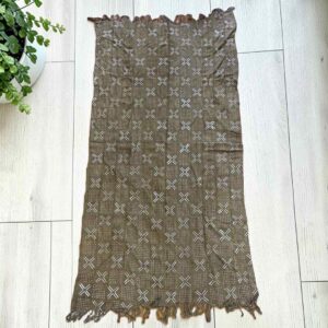 Medium muted earth-toned mudcloth with pale crosses and dots, laid on a timber surface with green leaves