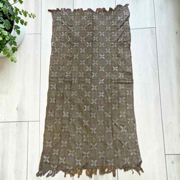 Medium muted earth-toned mudcloth with pale crosses and dots, laid on a timber surface with green leaves