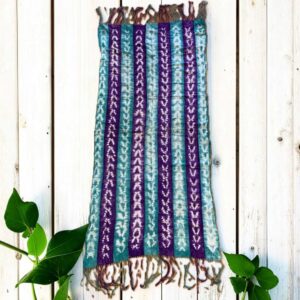 Small mudcloth with alternating jewel-toned strips and pale chevrons, laid on a timber surface with green leaves