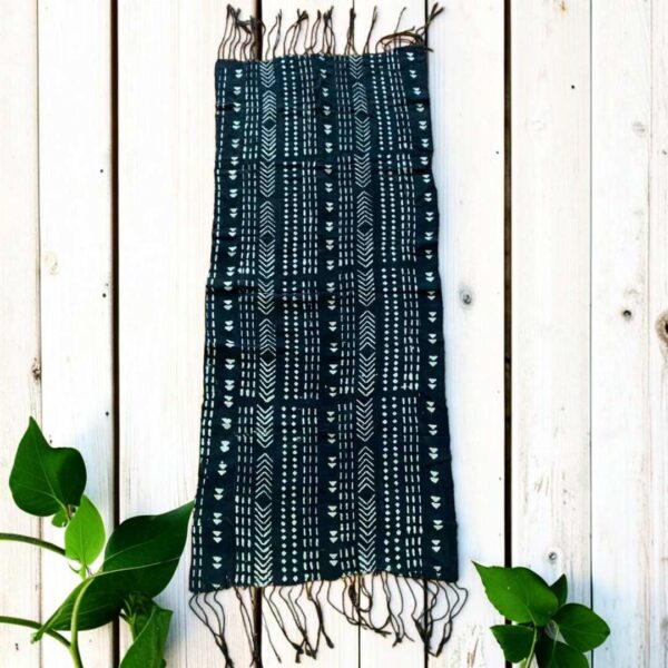 bashiri-mudcloth-small-166-harvest-african-textile-bogolan-main Small dark mudcloth with columns of pale chevrons and triangles, laid on a timber surface with green leaves