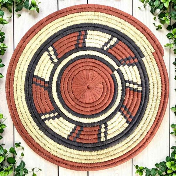bashiri-placemat-178-harmony-burkina-platter-woven-main Woven raffia rust, black and natural Burkina platter with a wheel pattern, on white timber with greenery