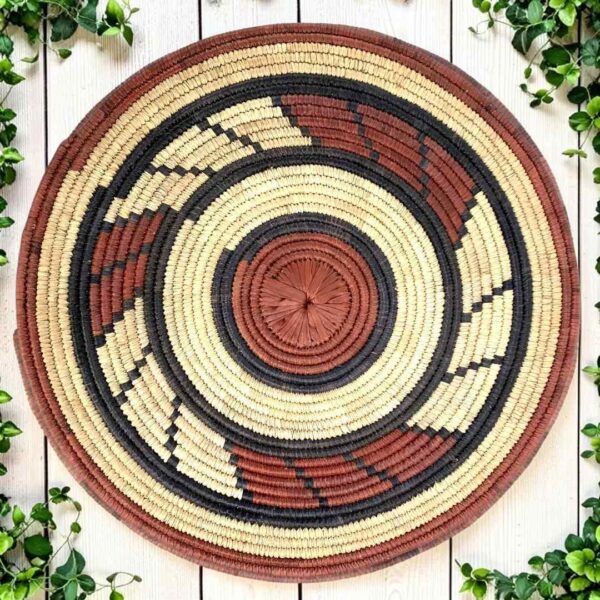 Woven raffia rust, black and natural Burkina platter with a swirl pattern, on white timber with greenery