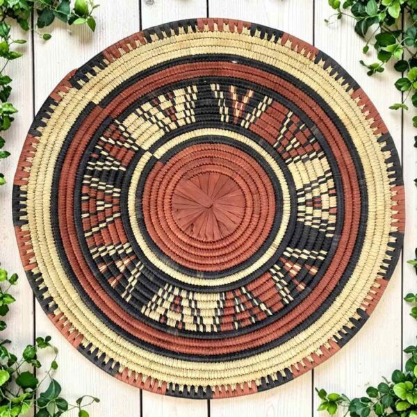 bashiri-placemat-182-sunbeam-burkina-platter-woven-main Woven raffia rust, black and natural Burkina platter with a starburst pattern, on white timber with greenery