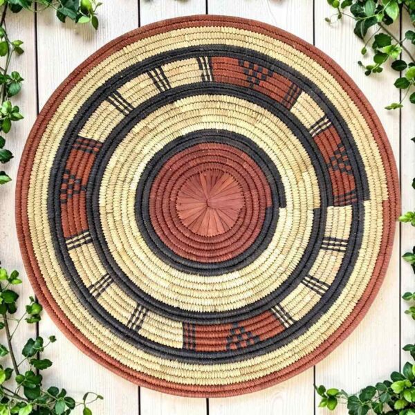 Woven raffia rust, black and natural Burkina platter with a concentric pattern, on white timber with greenery