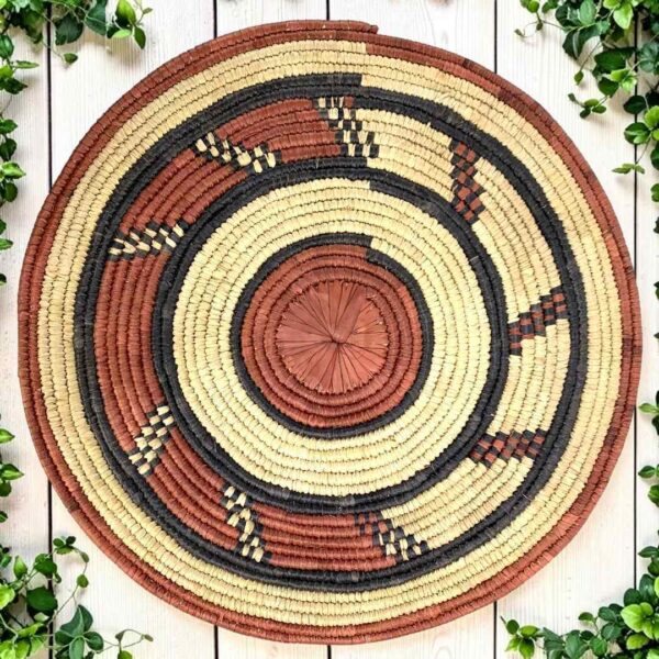 Woven raffia rust, black and natural Burkina platter with a hook pattern, on white timber with greenery
