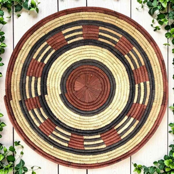 bashiri-placemat-185-tranquil-burkina-platter-handwoven-main Woven raffia rust, black and natural Burkina platter with a band pattern, on white timber with greenery