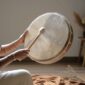 Hands playing a shamanic frame drum with a wooden mallet, styled in a warm meditation setting