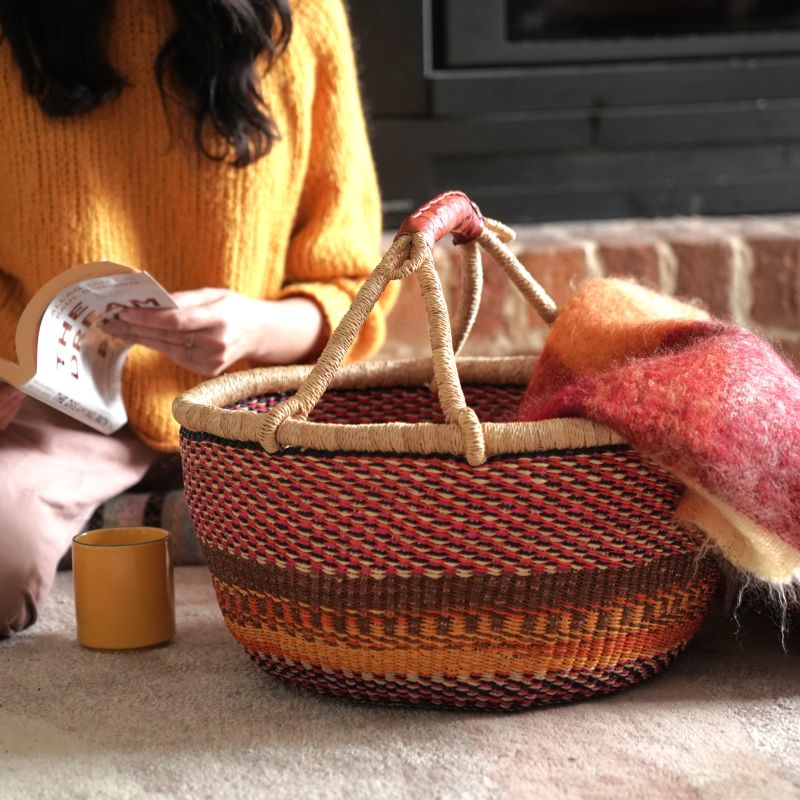 XL round Bolga basket in rust, brown and natural elephant grass weave with leather handle, styled in a home setting with a blanket inside, handwoven in Bolgatanga Ghana
