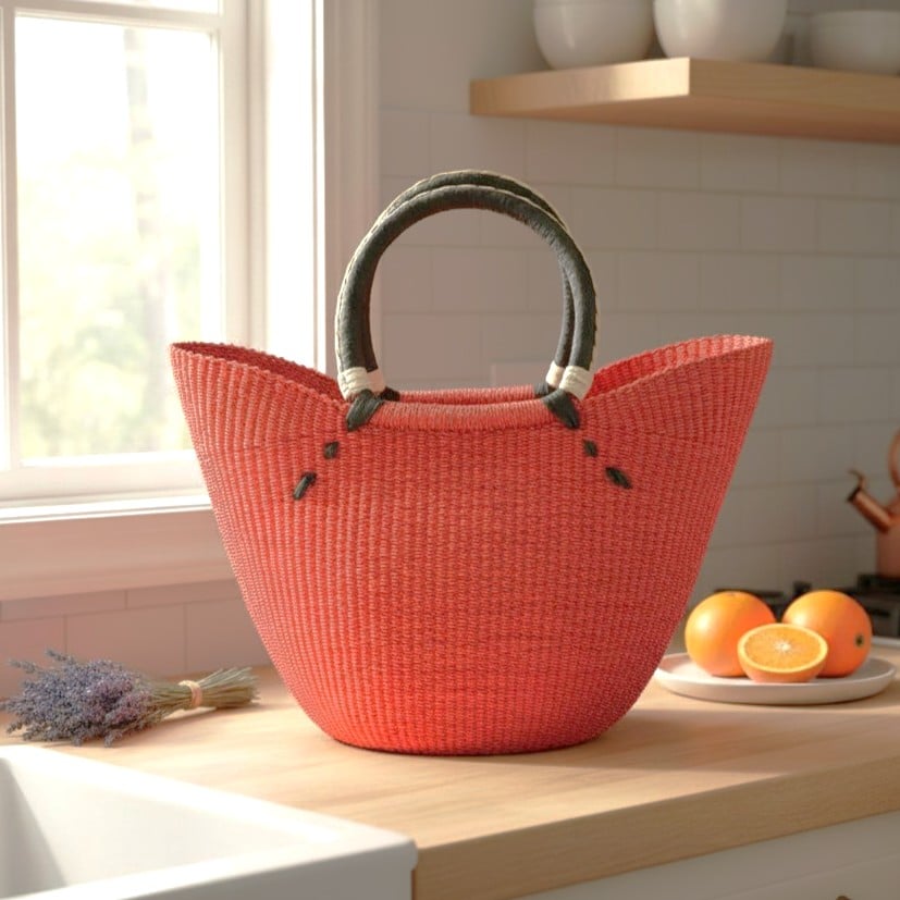 a woven market basket n a red colour with a black leather handle, positioned on a bright kitchen bench.