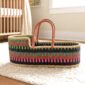 Moses baby basket with dark multicolour stripes and leather handles on a rug beside a cot