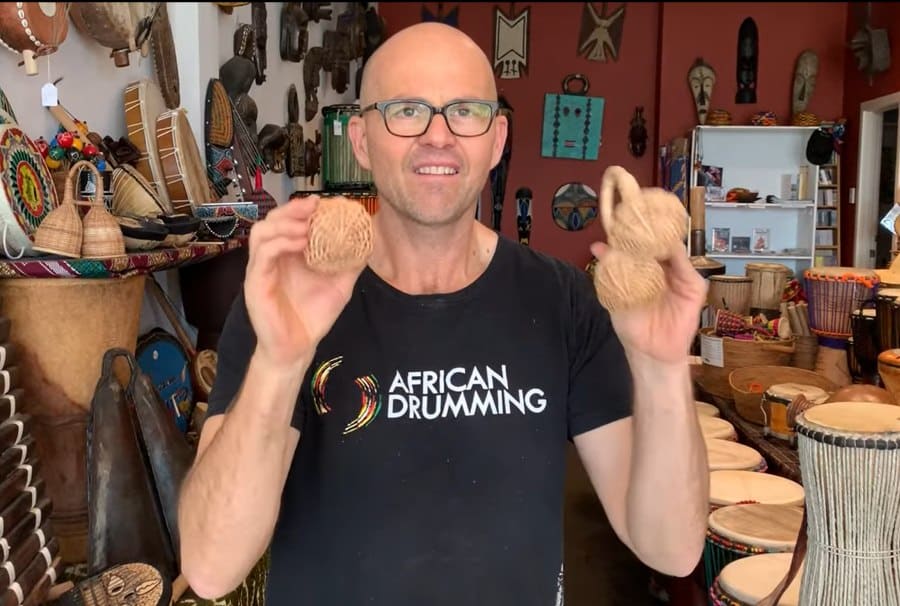 Simon holding two cane jinglers in the Bashiri store, surrounded by drums, masks, and percussion instruments