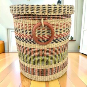 Closed laundry basket with natural straw with pink, green and black bands, a woven lid and leather ring handles