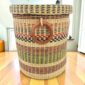 Closed laundry basket with natural straw with pink, green and black bands, a woven lid and leather ring handles