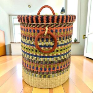 Closed laundry basket with multicoloured orange, blue and yellow stripes above a natural base, with a woven lid and leather ring handles