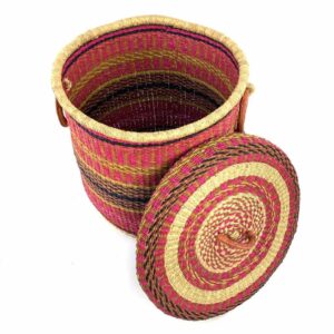 The closed laundry basket with its lid off, showing bright pink with gold and black bands, a patterned woven lid and leather ring handles