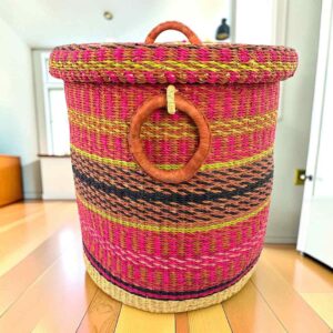 Closed laundry basket with bright pink with gold and black bands, a patterned woven lid and leather ring handles