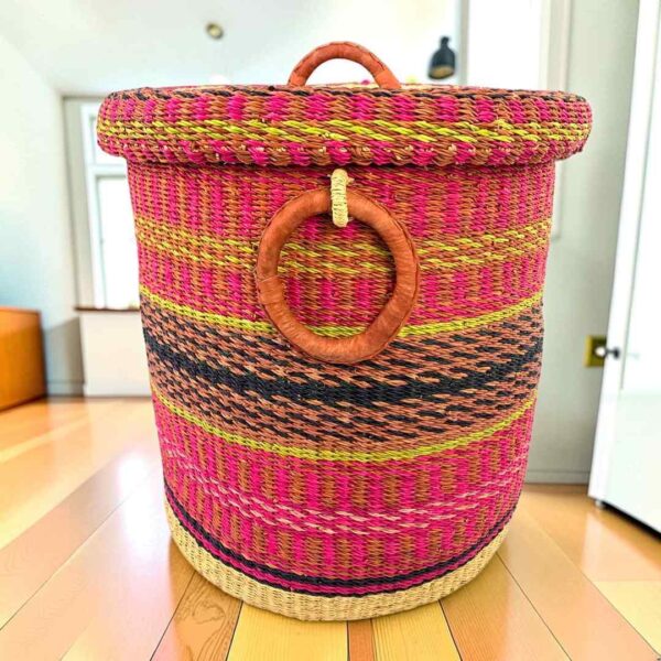 Closed laundry basket with bright pink with gold and black bands, a patterned woven lid and leather ring handles