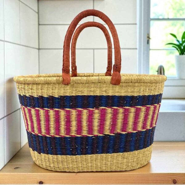 Extra large oval Bolga basket with navy bands framing a pink and cream striped band and leather handles, on a wooden kitchen bench