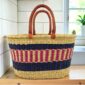 Extra large oval Bolga basket with navy bands framing a pink and cream striped band and leather handles, on a wooden kitchen bench
